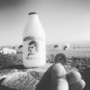 Black & white picture of a hand holding a coin up beside a glass pint bottle of Mossgiel Organic Milk.